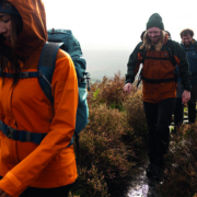 Vrouw en twee mannen wandelen door de natte, heuvelachtige bergen in herfst/winter kleding, met mutsen en wandelrugzakken. De vrouw draagt een oranje jas, man 1 een bruine en zwarte jas, en man 2 een donkerblauwe jas. Het pad is nat en de omgeving is grijs en grauw