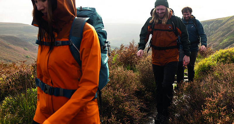 Vrouw en twee mannen wandelen door de natte, heuvelachtige bergen in herfst/winter kleding, met mutsen en wandelrugzakken. De vrouw draagt een oranje jas, man 1 een bruine en zwarte jas, en man 2 een donkerblauwe jas. Het pad is nat en de omgeving is grijs en grauw